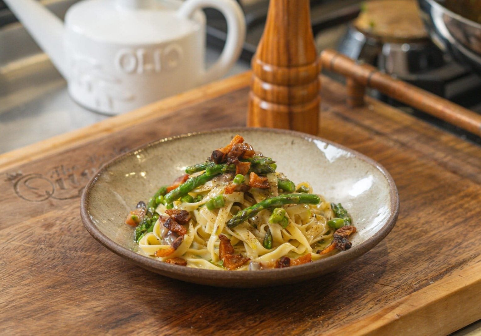 A bowl of pasta with green vegetables and diced meat on a wooden cutting board, set beside a wooden pepper grinder and a white kettle.