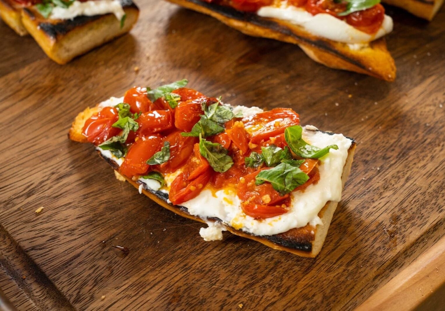 Toasted bread topped with melted cheese, roasted tomatoes, and fresh basil on a wooden surface.