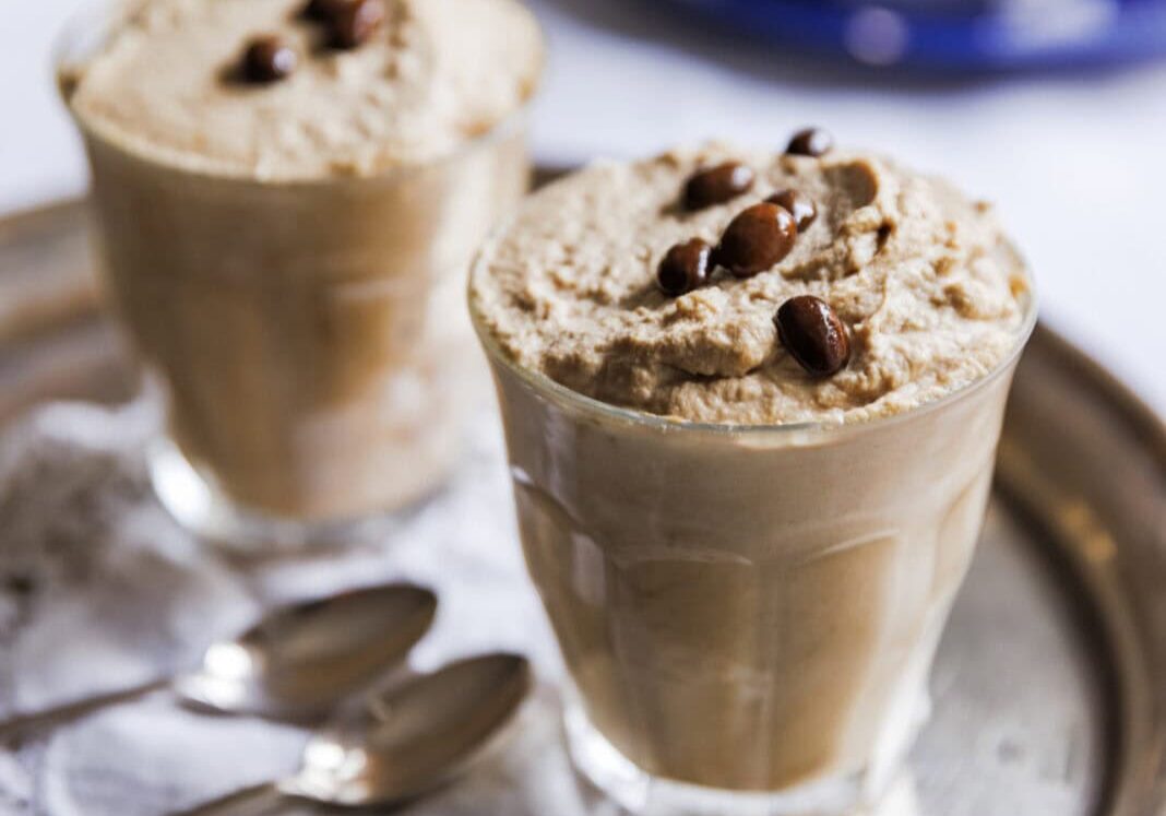 Two glasses of whipped coffee topped with coffee beans on a silver tray, accompanied by two spoons on a lace doily.