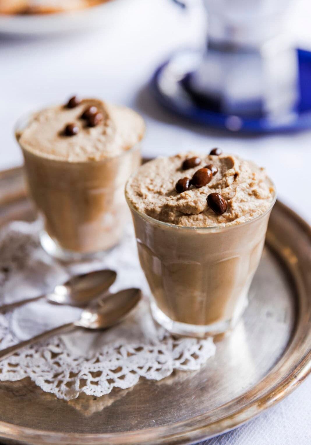 Two glasses of whipped coffee topped with coffee beans on a silver tray, accompanied by two spoons on a lace doily.