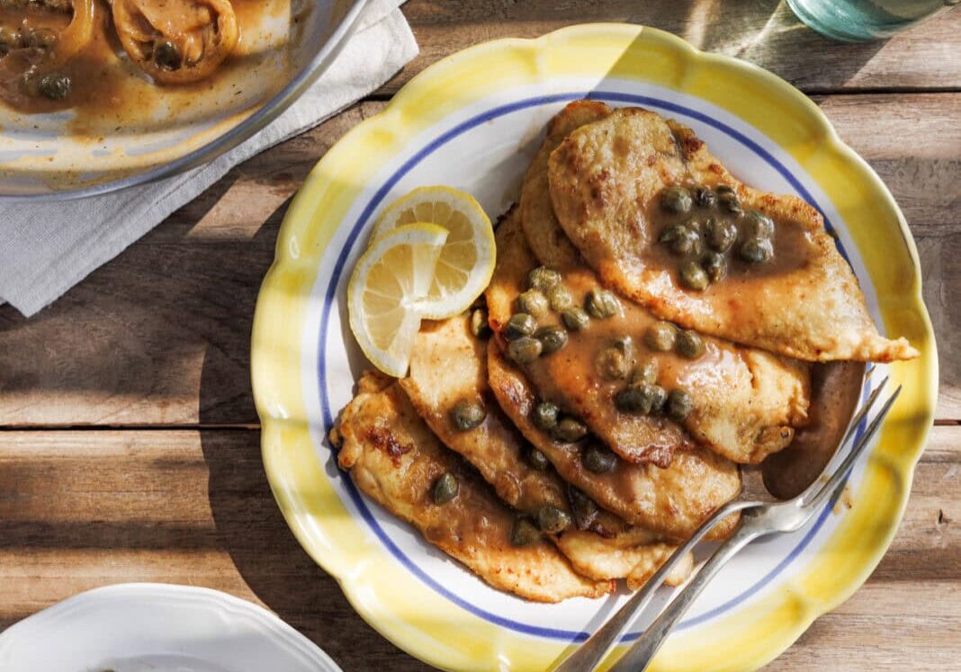Chicken piccata with capers on a yellow-rimmed plate, accompanied by a lemon slice. Nearby are a serving dish, a smaller plate with a piece, and two green glass bottles on a wooden table.