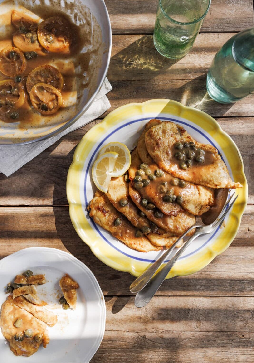 Chicken piccata with capers on a yellow-rimmed plate, accompanied by a lemon slice. Nearby are a serving dish, a smaller plate with a piece, and two green glass bottles on a wooden table.