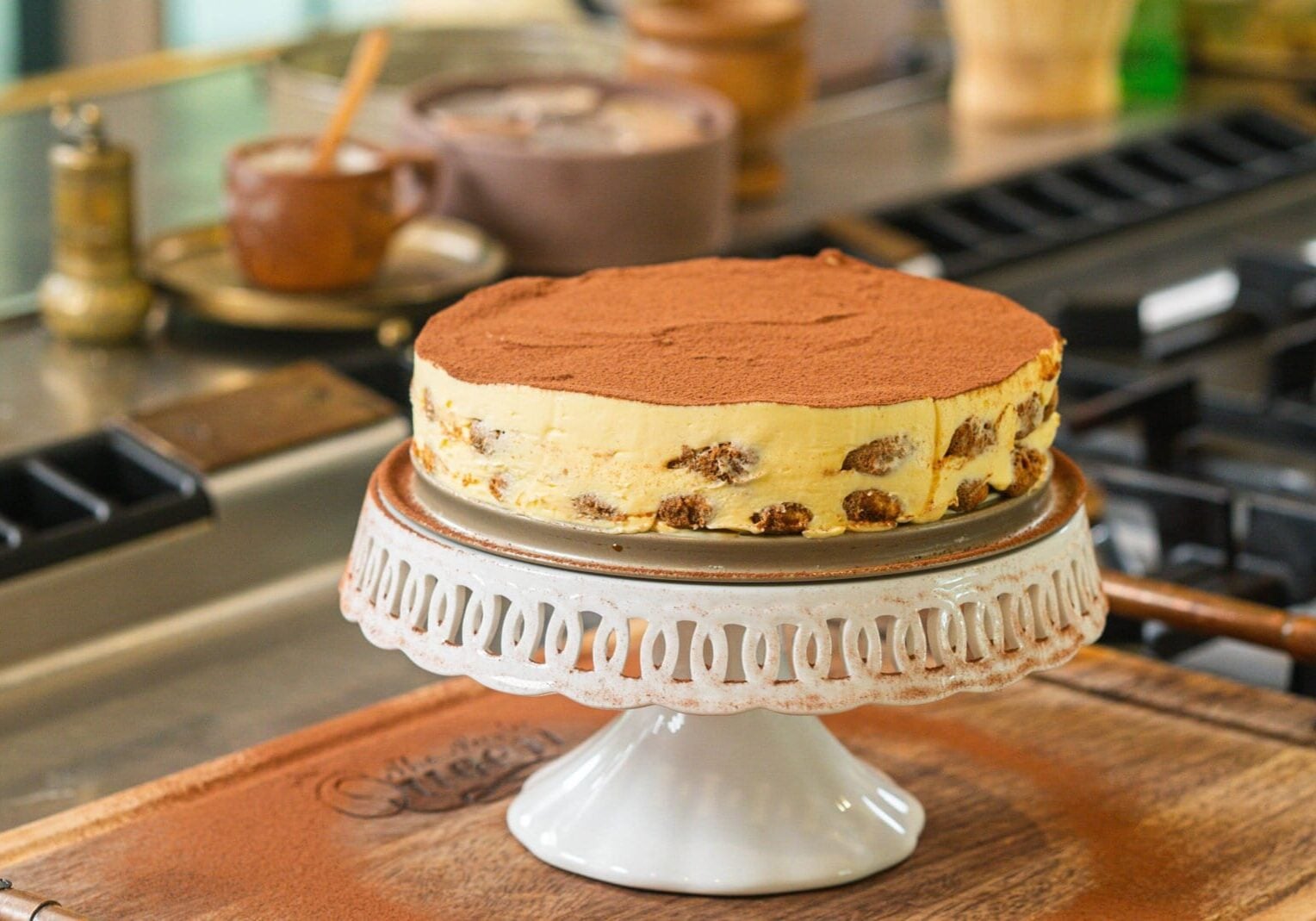 Tiramisu topped with cocoa powder on a white cake stand, placed on a wooden board in a kitchen setting.
