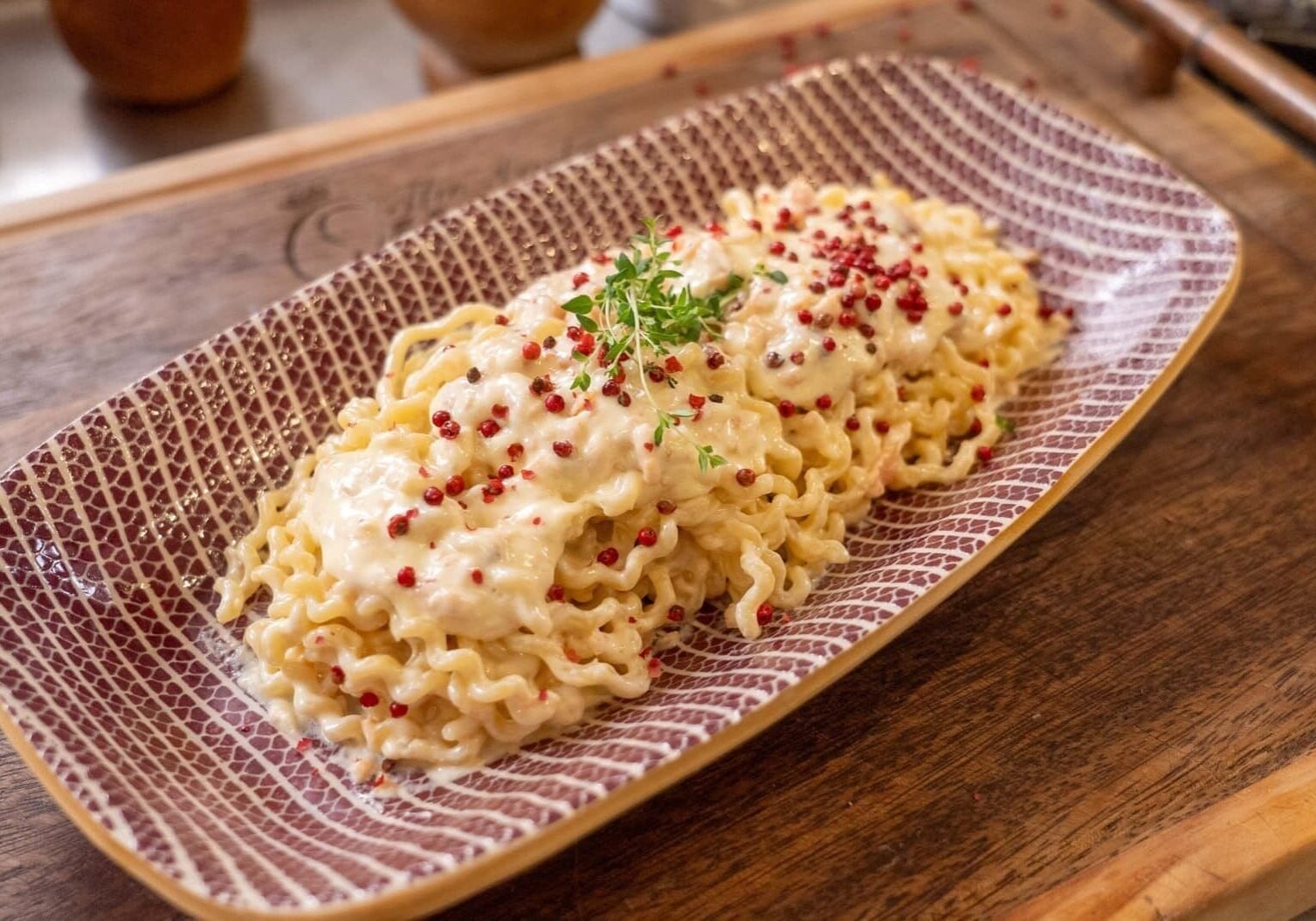 A rectangular plate with pasta topped with creamy sauce, pink peppercorns, and garnished with fresh greens, placed on a wooden surface.