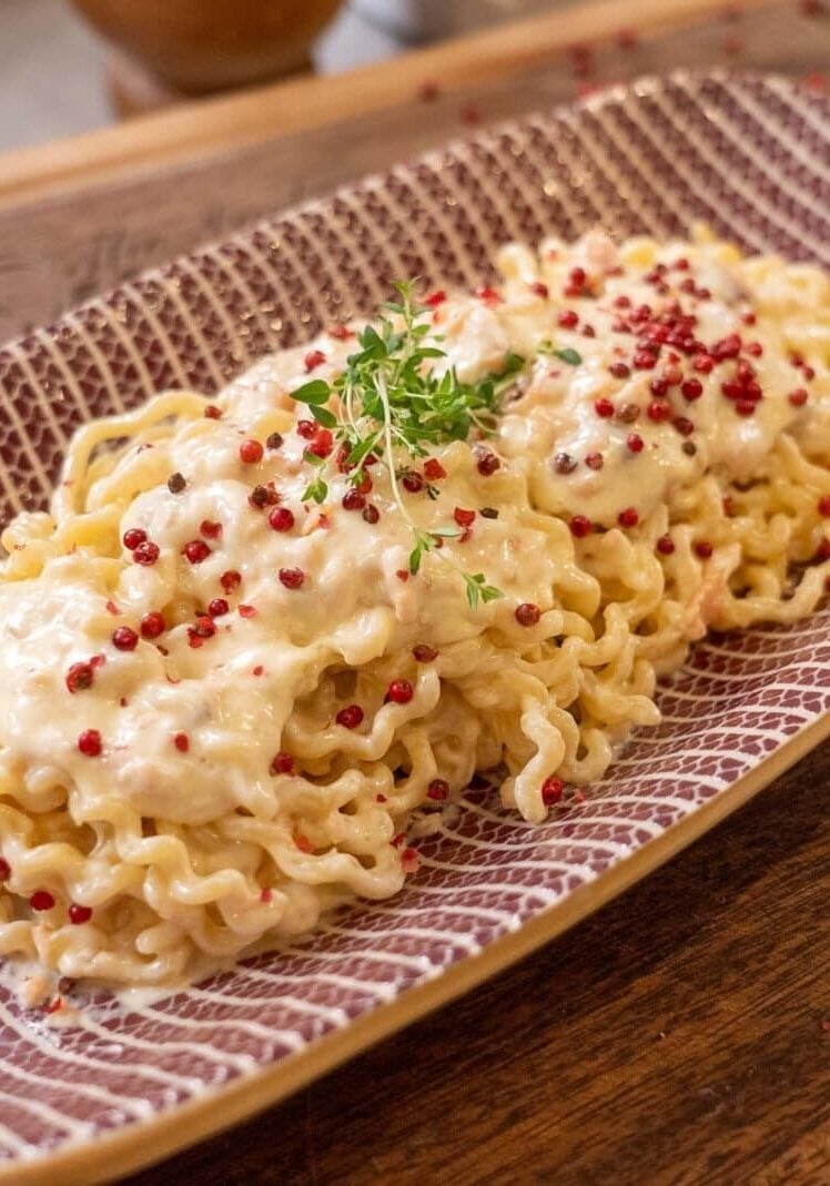 A rectangular plate with pasta topped with creamy sauce, pink peppercorns, and garnished with fresh greens, placed on a wooden surface.