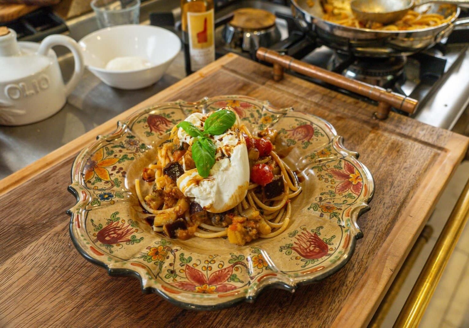 A plate of pasta topped with a ball of burrata, roasted vegetables, and a basil leaf, served on a floral-patterned dish on a wooden cutting board.