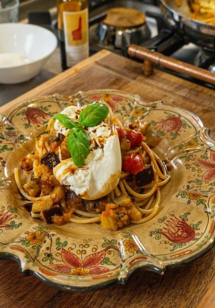 A plate of pasta topped with a ball of burrata, roasted vegetables, and a basil leaf, served on a floral-patterned dish on a wooden cutting board.