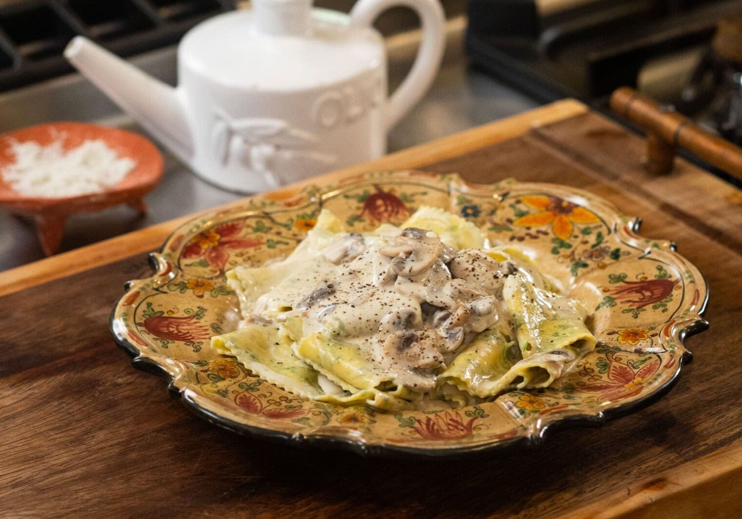A plate of ravioli topped with creamy mushroom sauce sits on a decorative dish, with a teapot and grated cheese in the background on a wooden surface.