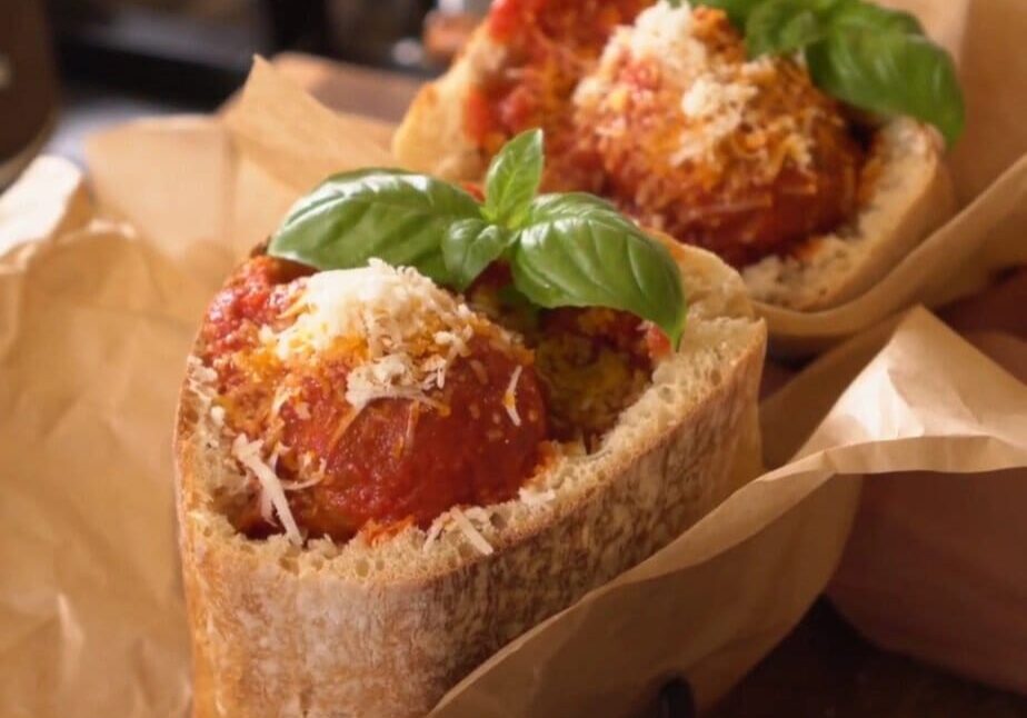 Two meatball subs in crusty bread with tomato sauce, grated cheese, and basil leaves, wrapped in parchment paper.