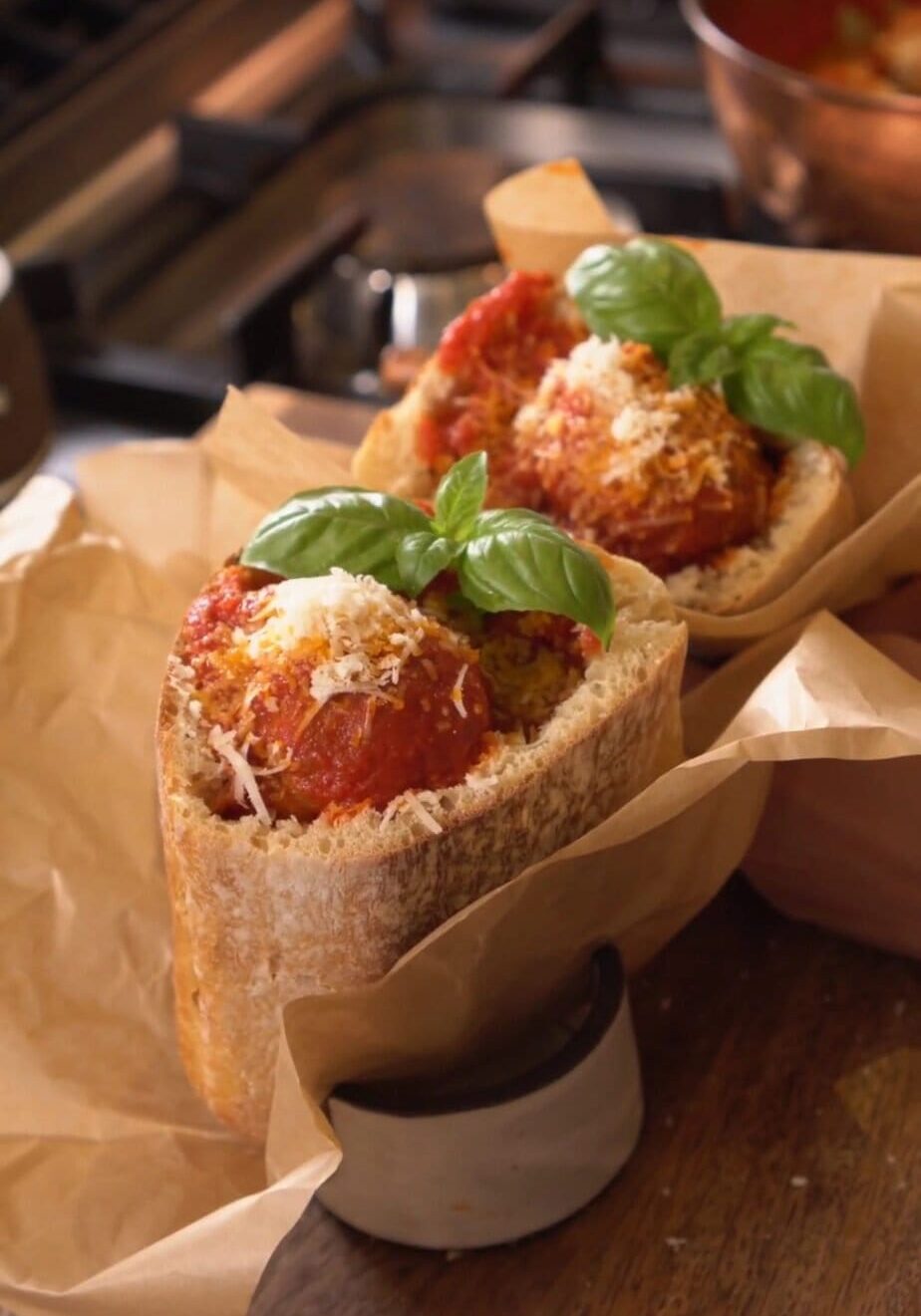 Two meatball subs in crusty bread with tomato sauce, grated cheese, and basil leaves, wrapped in parchment paper.