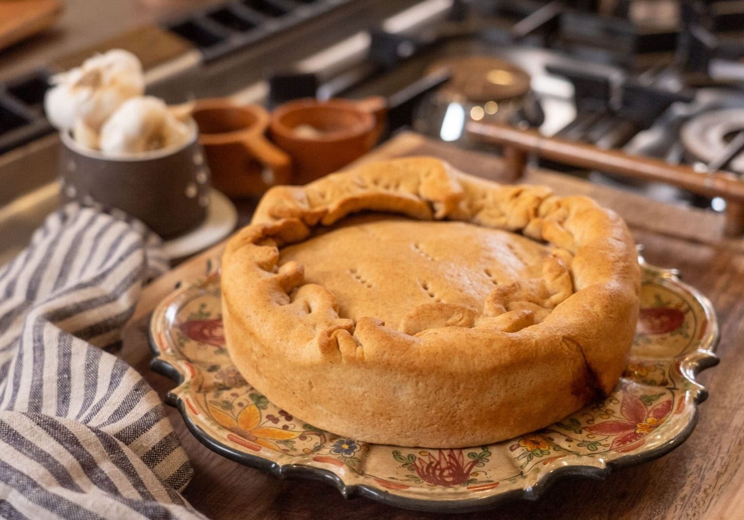 A round, golden-brown pie sits on a decorative plate beside a striped kitchen towel on a wooden surface near a stove.