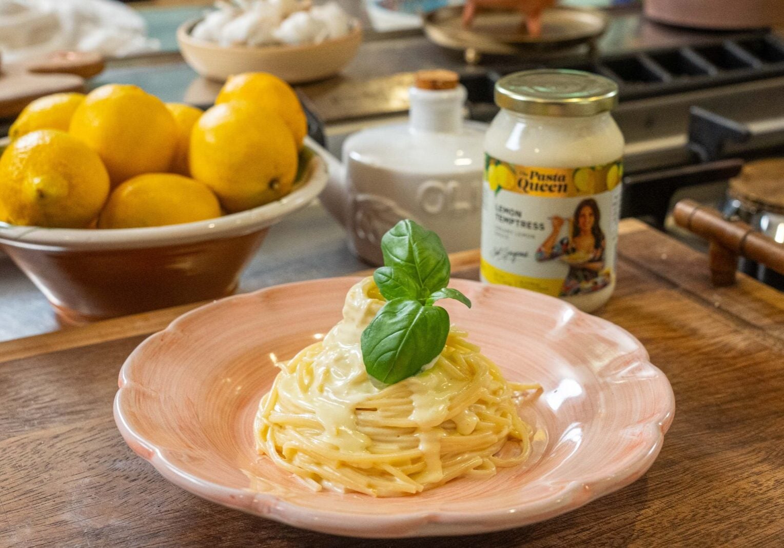 A plate of creamy pasta garnished with basil is set on a wooden counter beside a bowl of lemons and a jar of lemon alfredo sauce.