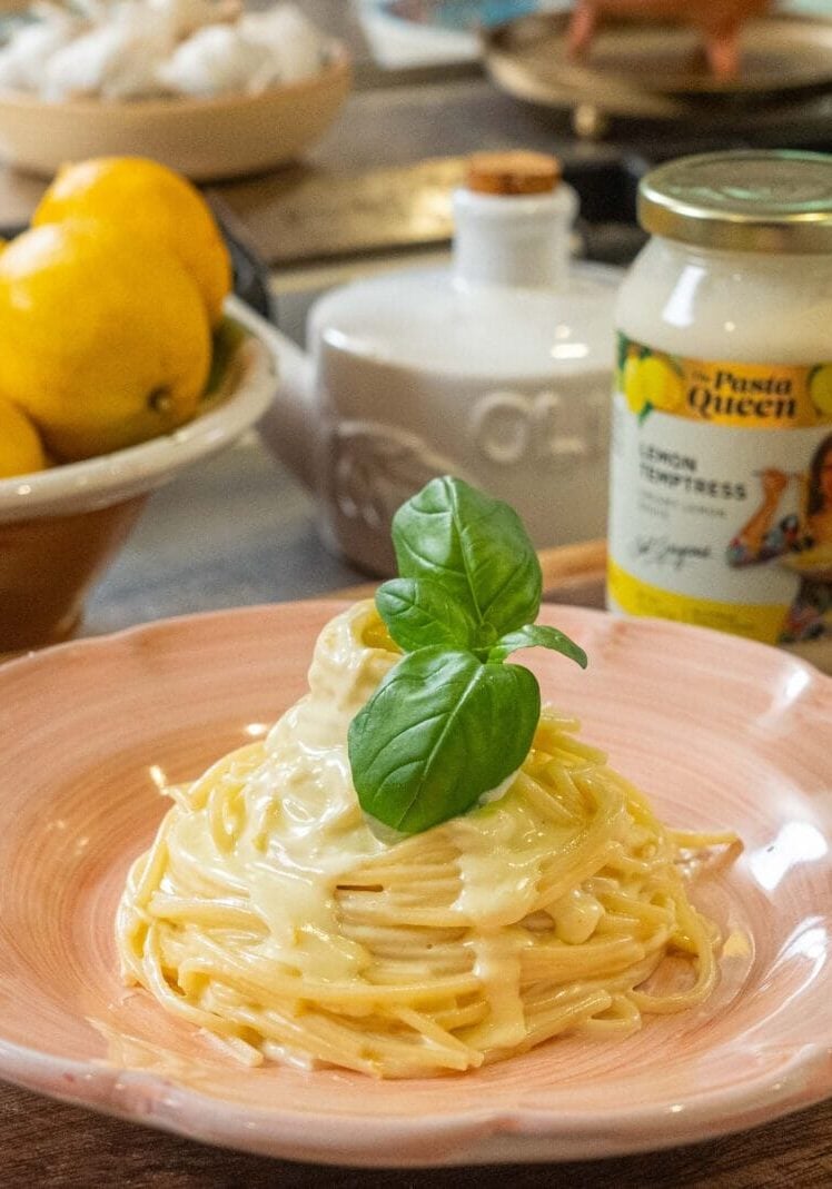 A plate of creamy pasta garnished with basil is set on a wooden counter beside a bowl of lemons and a jar of lemon alfredo sauce.