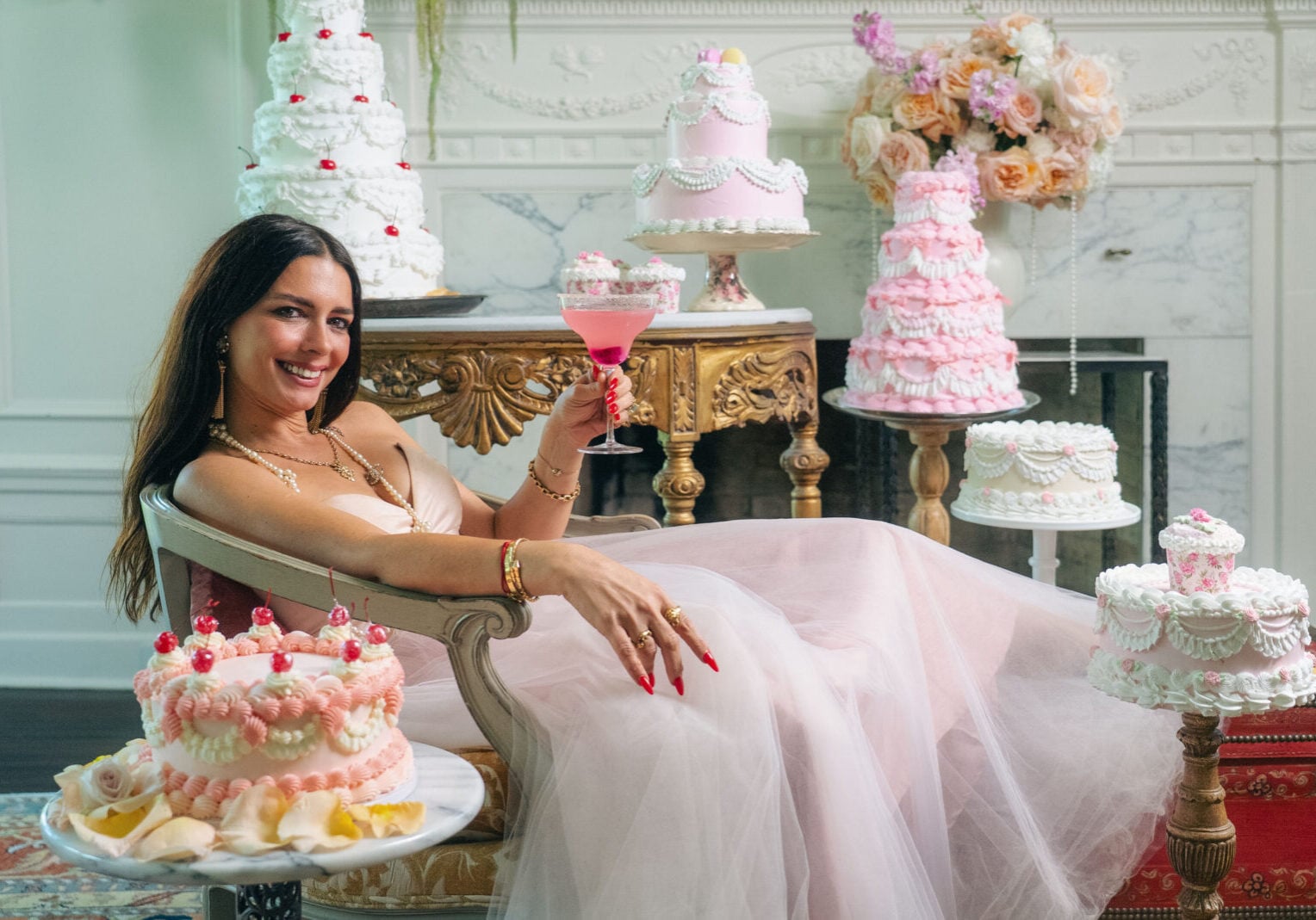 A woman in a pink dress sits on a chaise lounge, holding a pink drink, surrounded by ornate, pastel-colored cakes in an elegant room.