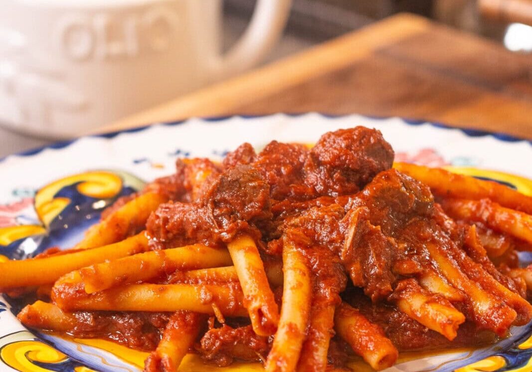A plate of pasta topped with a rich tomato and meat sauce sits on a colorful ceramic dish on a wooden surface, with an olive oil container in the background.