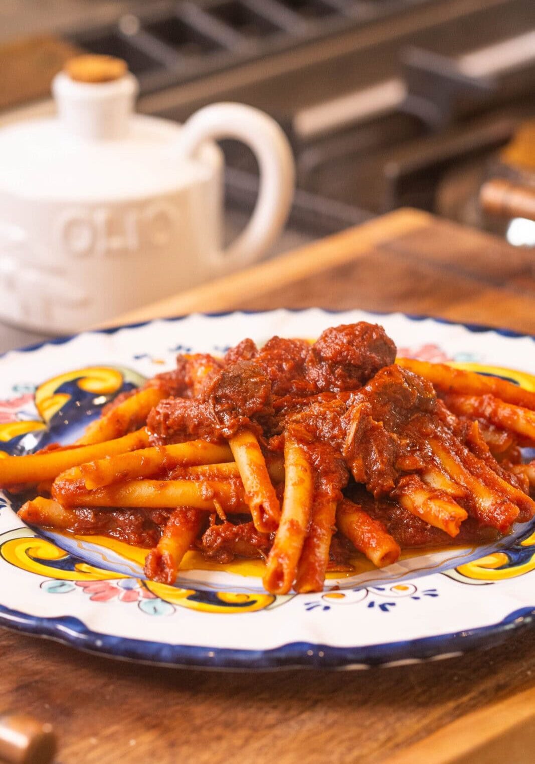 A plate of pasta topped with a rich tomato and meat sauce sits on a colorful ceramic dish on a wooden surface, with an olive oil container in the background.