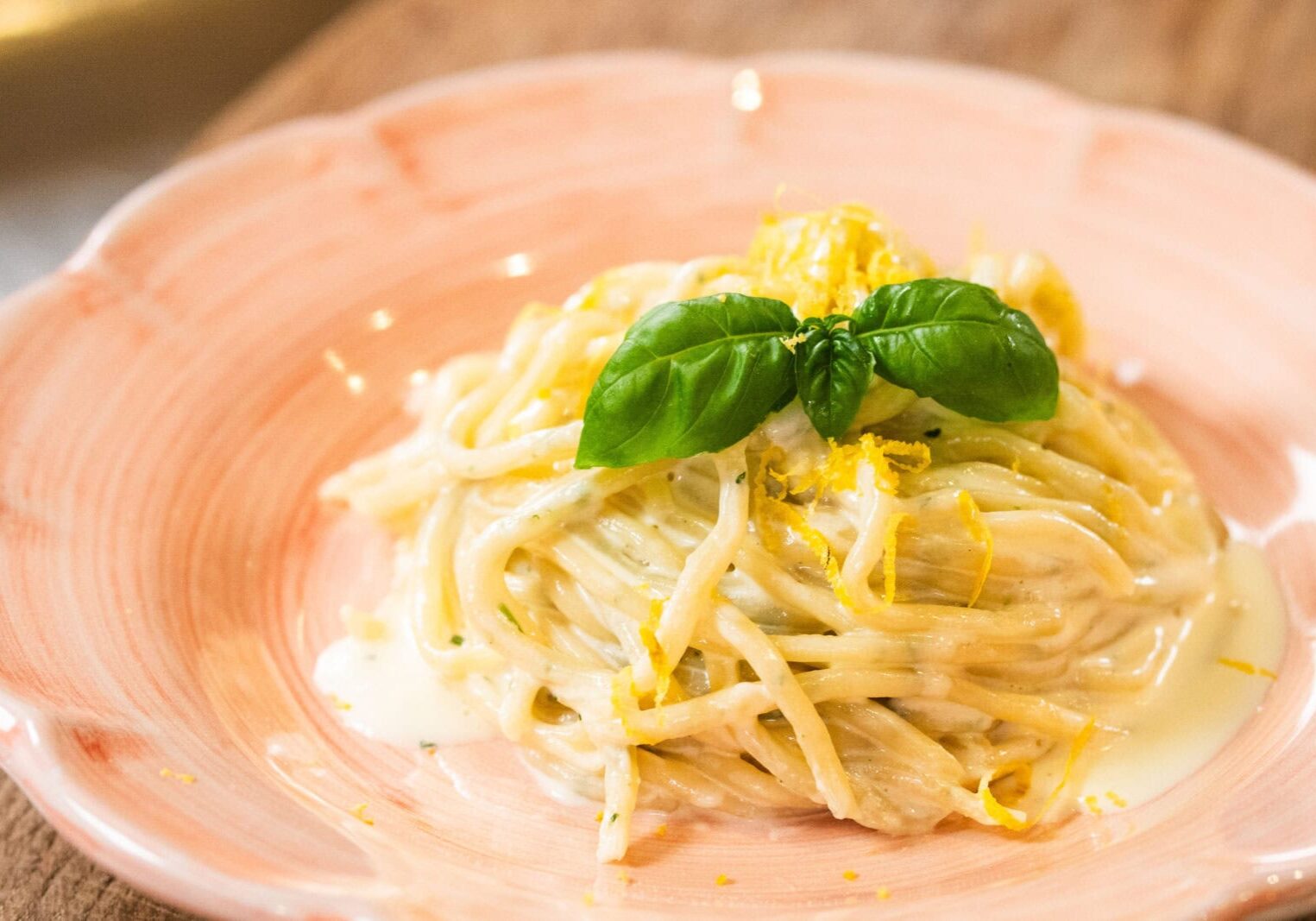 A plate of creamy spaghetti garnished with fresh basil leaves and lemon zest, served on a pink ceramic dish.