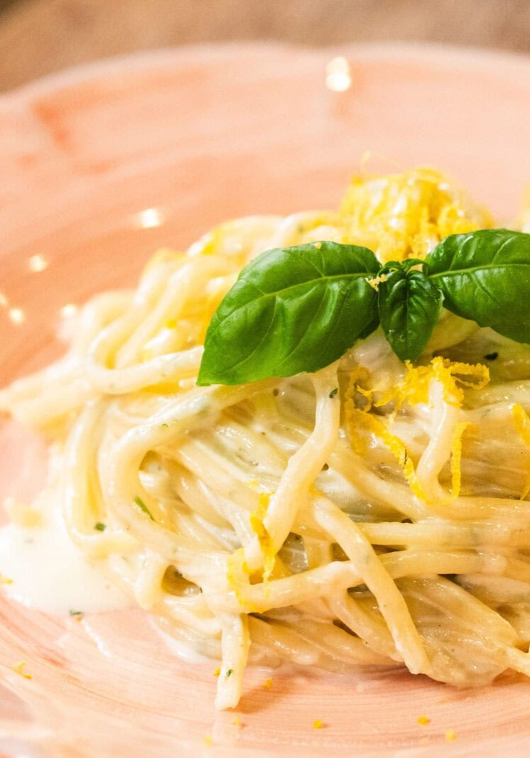 A plate of creamy spaghetti garnished with fresh basil leaves and lemon zest, served on a pink ceramic dish.