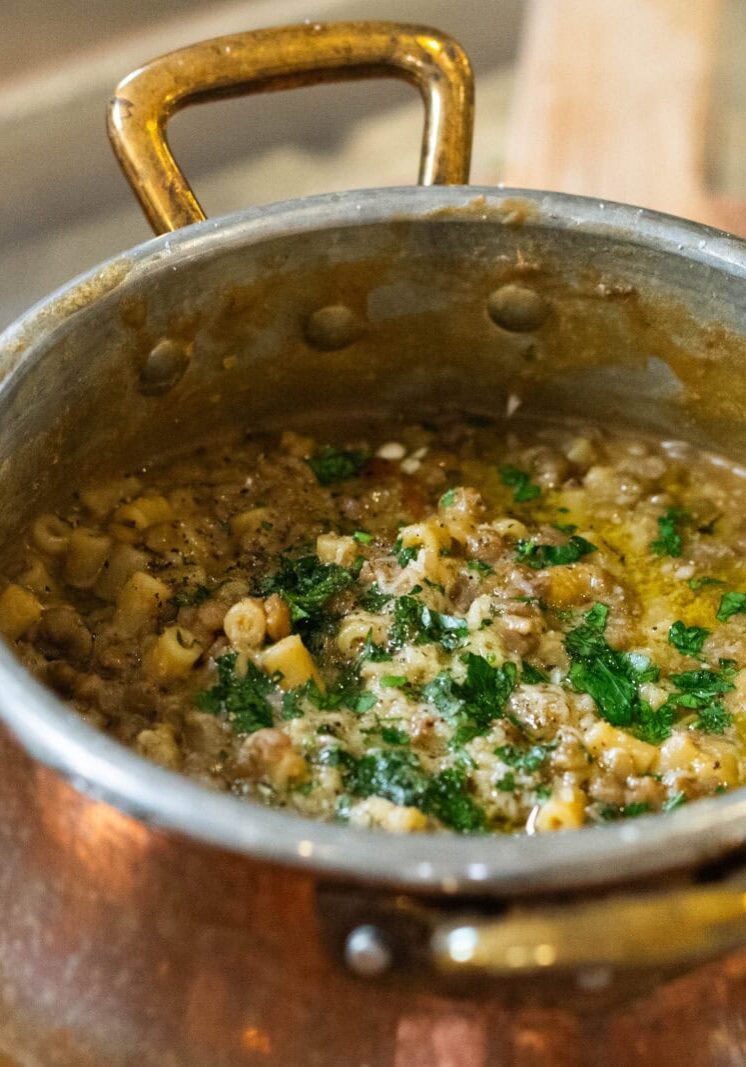 A copper pot filled with a thick, chunky soup or stew containing pasta, herbs, and possibly lentils or minced meat, placed on a wooden surface.