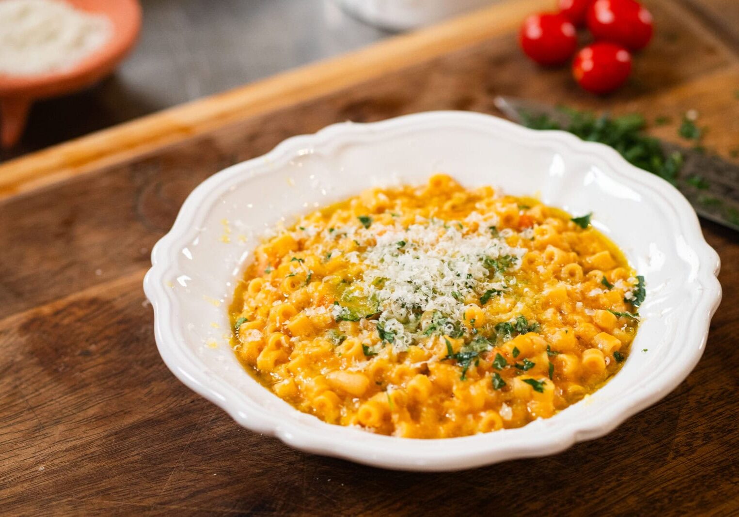 A white bowl filled with pasta in a creamy orange sauce, topped with grated cheese and chopped herbs, placed on a wooden surface.