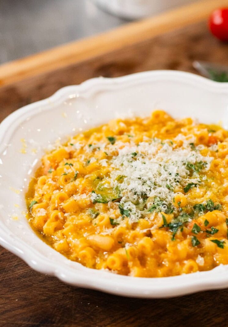 A white bowl filled with pasta in a creamy orange sauce, topped with grated cheese and chopped herbs, placed on a wooden surface.