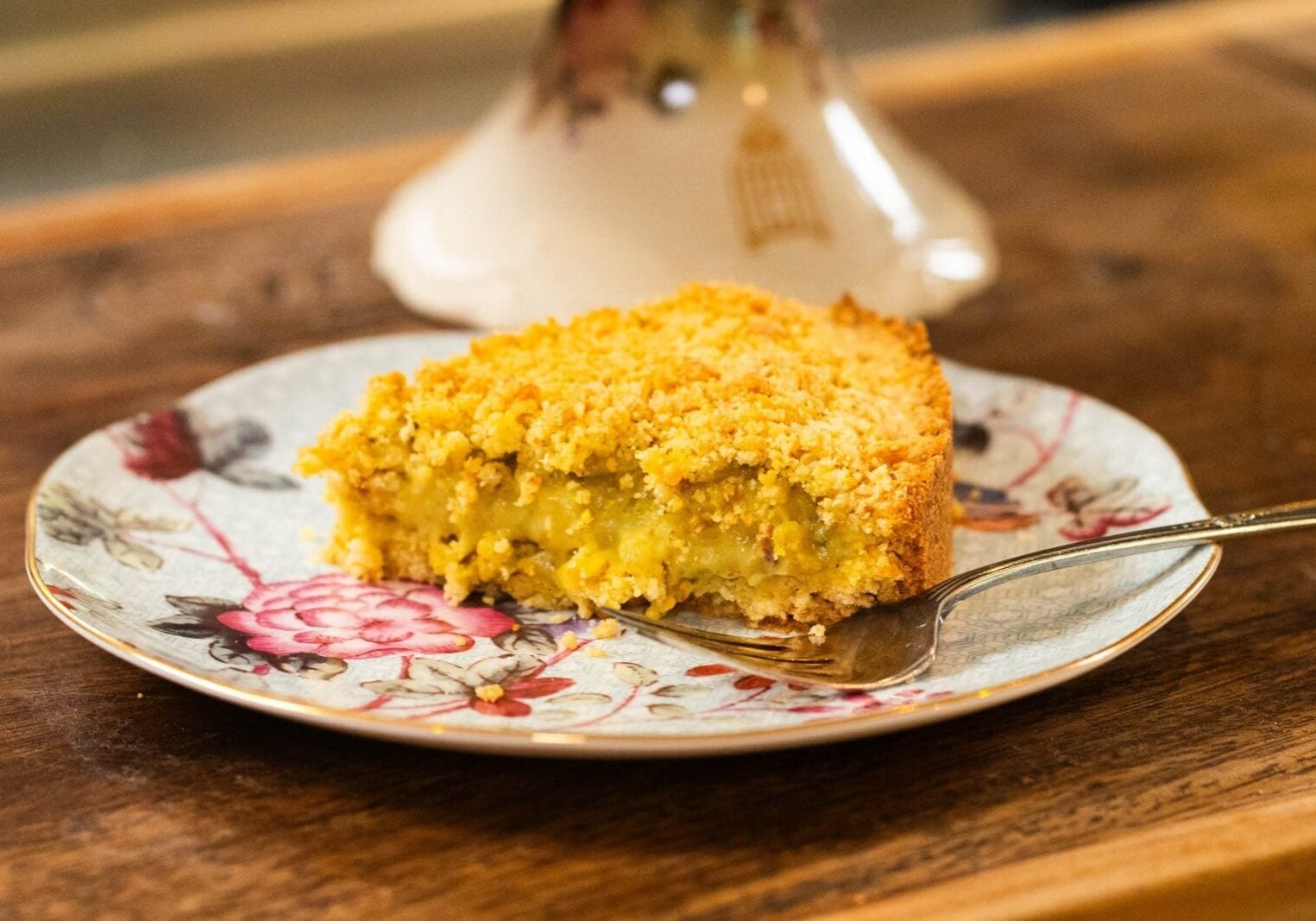 A slice of crumb-topped pie with a yellow filling is served on a floral-patterned plate with a fork on a wooden table.