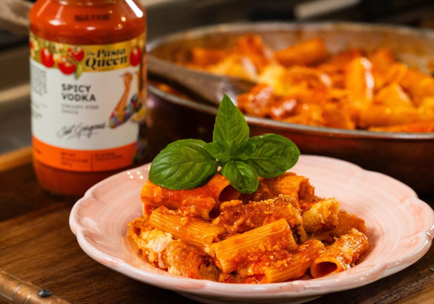 A plate of baked rigatoni pasta with tomato sauce and a basil garnish, next to a jar labeled "Spicy Vodka" pasta sauce and a pan of pasta in the background.