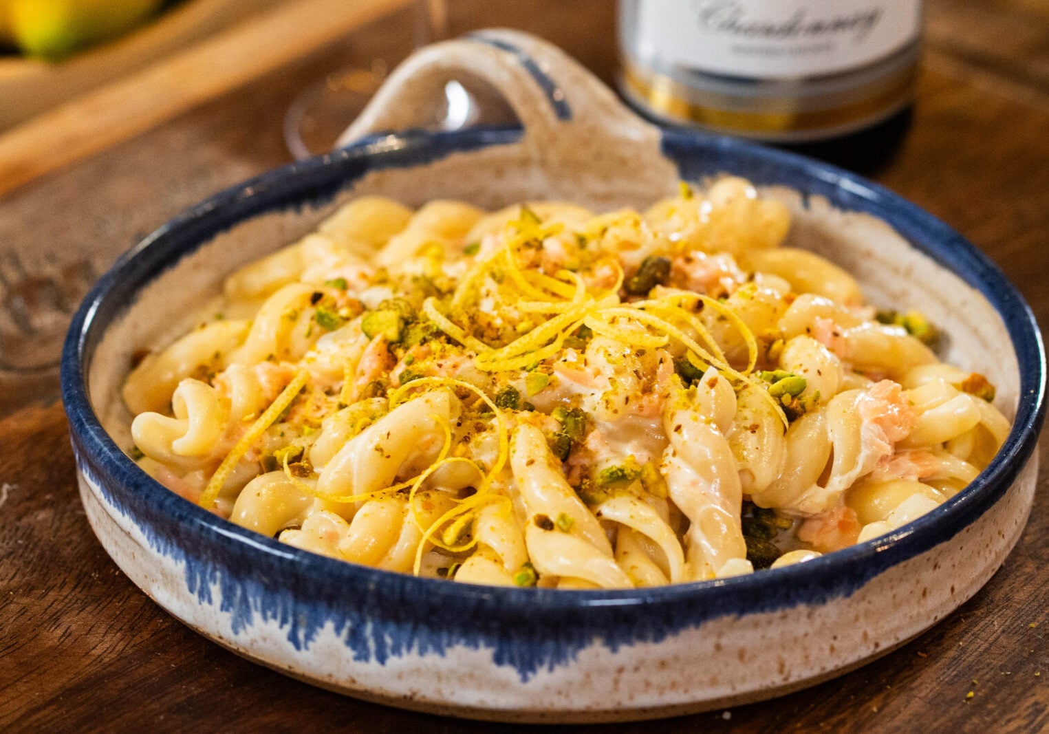 A ceramic dish of creamy pasta topped with lemon zest and chopped pistachios sits on a wooden table, with a bottle of white wine in the background.
