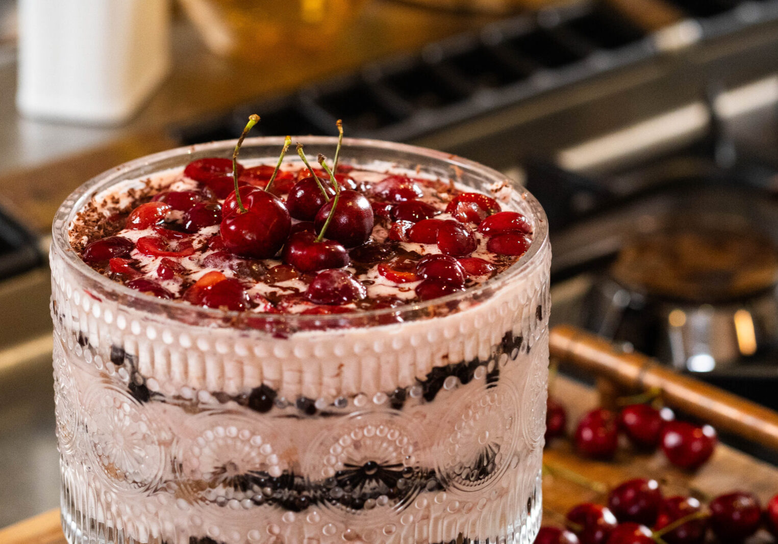 A glass trifle dish filled with layers of cream, chocolate, and topped with fresh cherries, sits on a kitchen counter near a stove.
