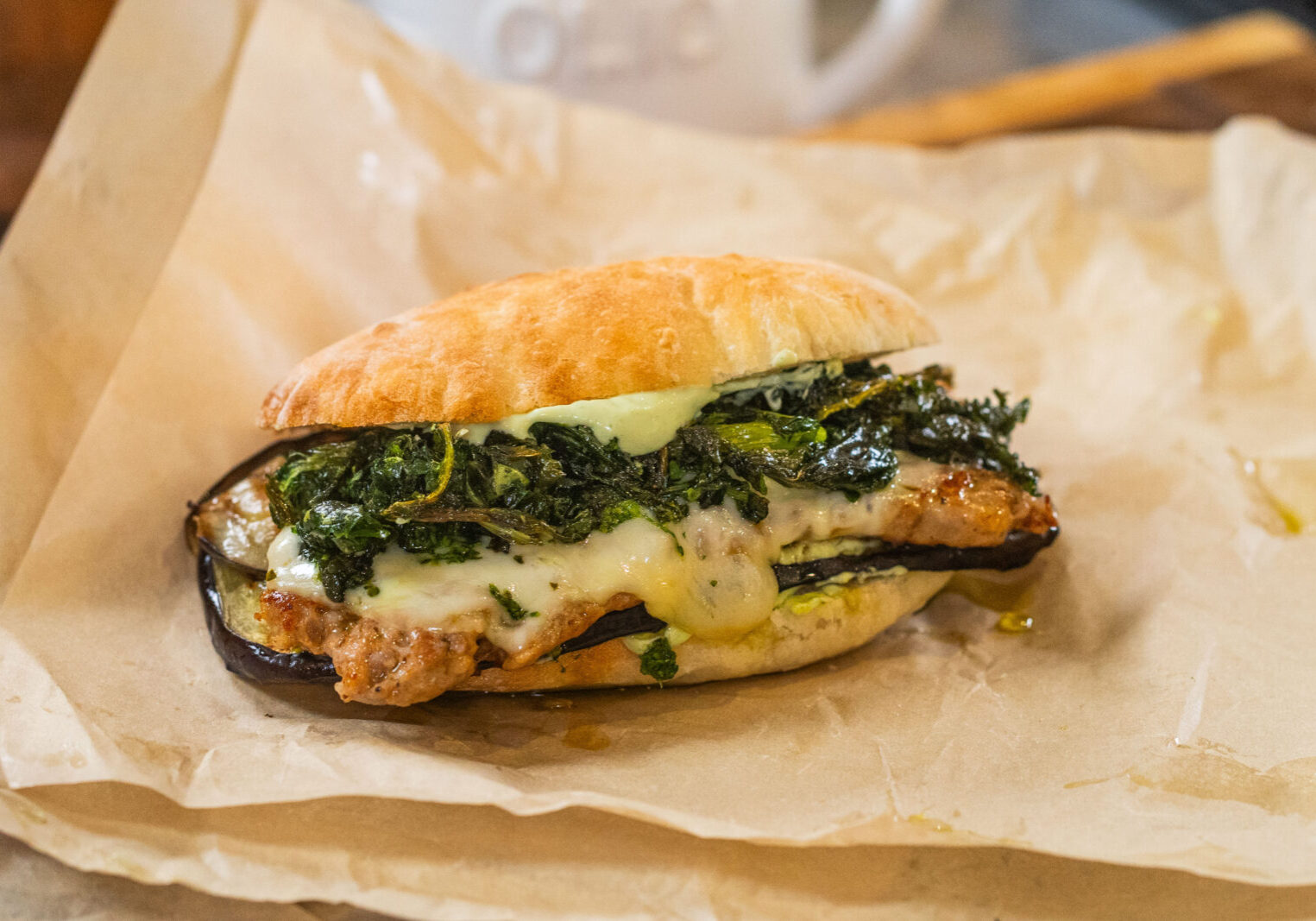 A sandwich on parchment paper filled with grilled eggplant, melted cheese, greens, and a slice of meat, with some oil visible on the bread and paper.