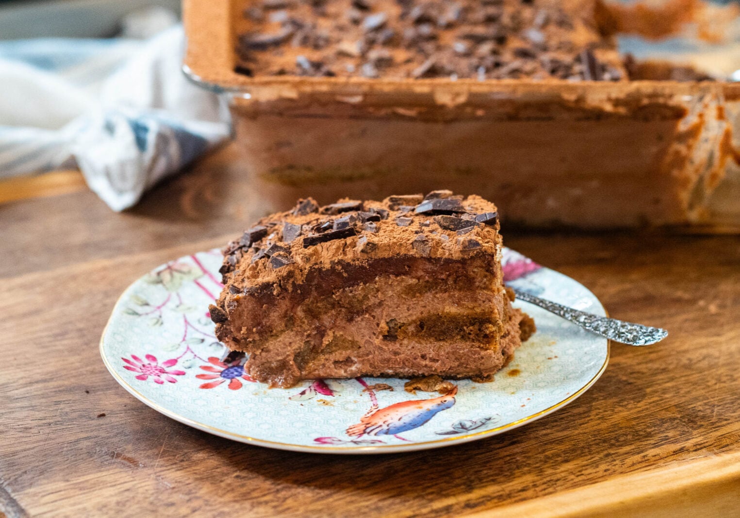 A slice of chocolate tiramisu with chocolate shavings on a decorative plate, with the rest of the dessert in a glass dish in the background.