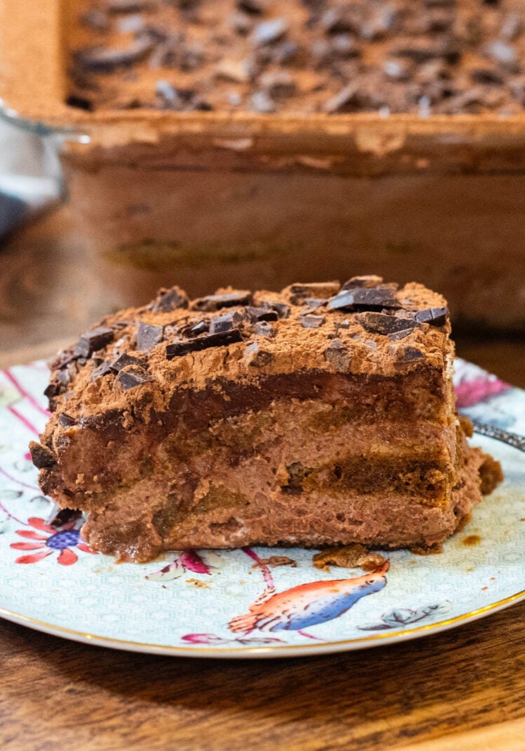 A slice of chocolate tiramisu with chocolate shavings on a decorative plate, with the rest of the dessert in a glass dish in the background.