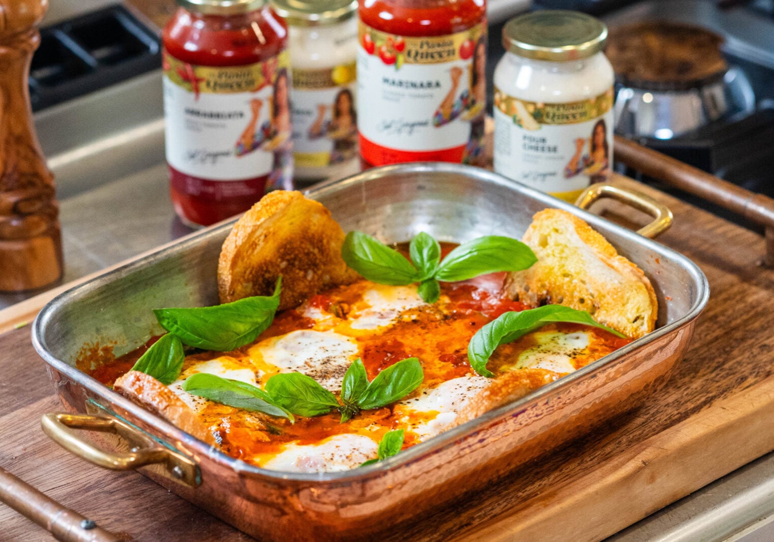 A copper baking dish with baked eggs in tomato sauce, topped with basil leaves and served with toasted bread, sits on a wooden board. Jars of pasta sauce are visible in the background.