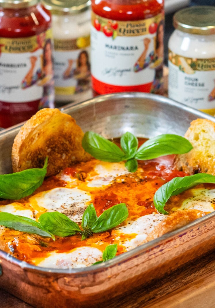 A copper baking dish with baked eggs in tomato sauce, topped with basil leaves and served with toasted bread, sits on a wooden board. Jars of pasta sauce are visible in the background.