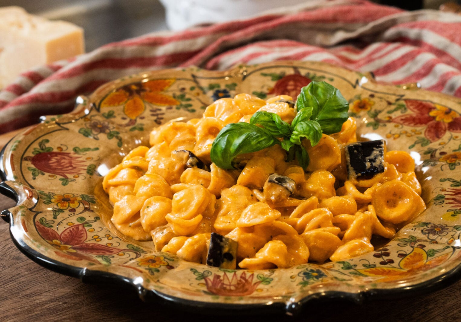 A decorative plate filled with orecchiette pasta in a creamy orange sauce, topped with basil leaves and small pieces of eggplant. A striped cloth and cheese block are in the background.