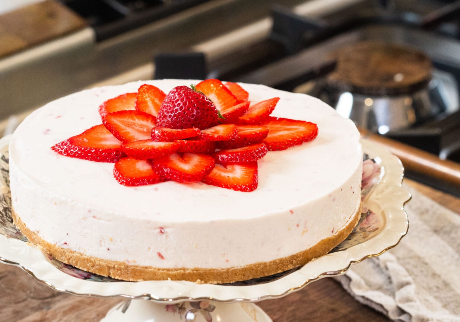 A cheesecake with a graham cracker crust, topped with sliced strawberries and a whole strawberry in the center, displayed on a floral cake stand in a kitchen setting.
