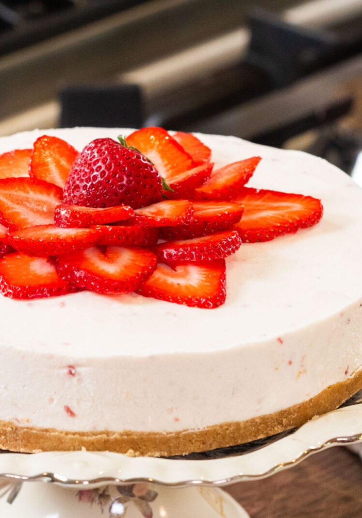 A cheesecake with a graham cracker crust, topped with sliced strawberries and a whole strawberry in the center, displayed on a floral cake stand in a kitchen setting.