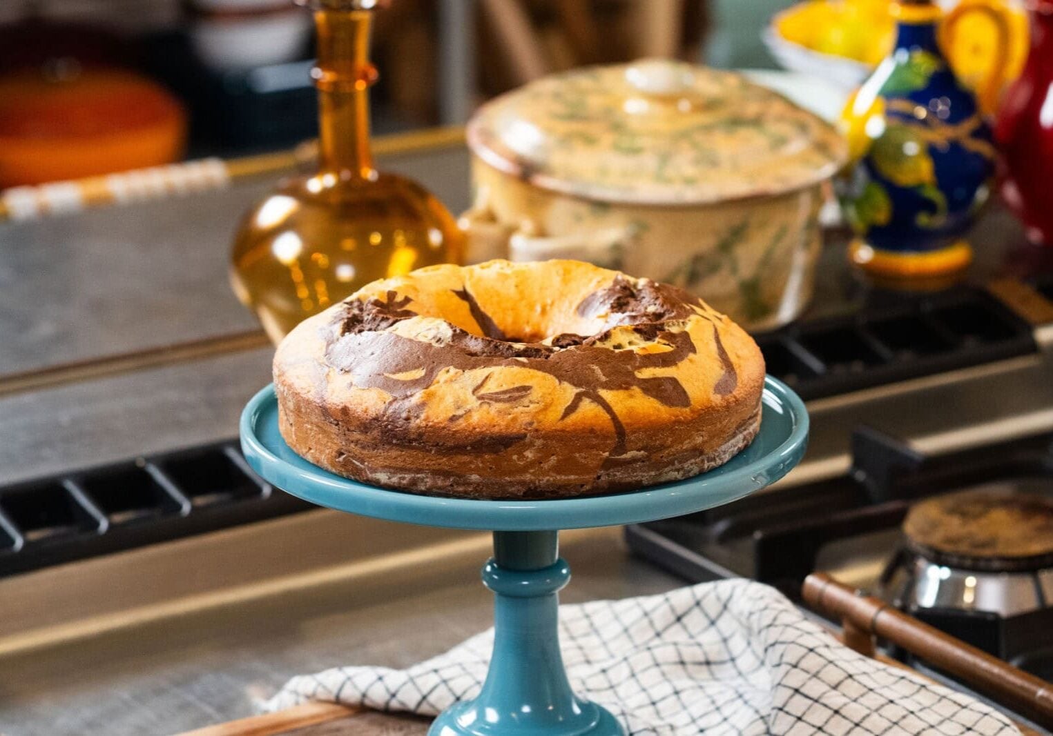 A marble cake with swirled chocolate and vanilla patterns sits on a blue cake stand in a kitchen setting.