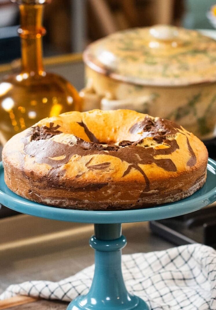 A marble cake with swirled chocolate and vanilla patterns sits on a blue cake stand in a kitchen setting.