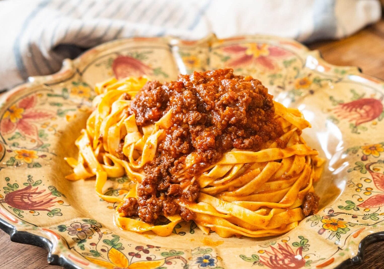 A plate of tagliatelle pasta topped with meat ragù sauce, served on a decorative floral dish with a cloth in the background.