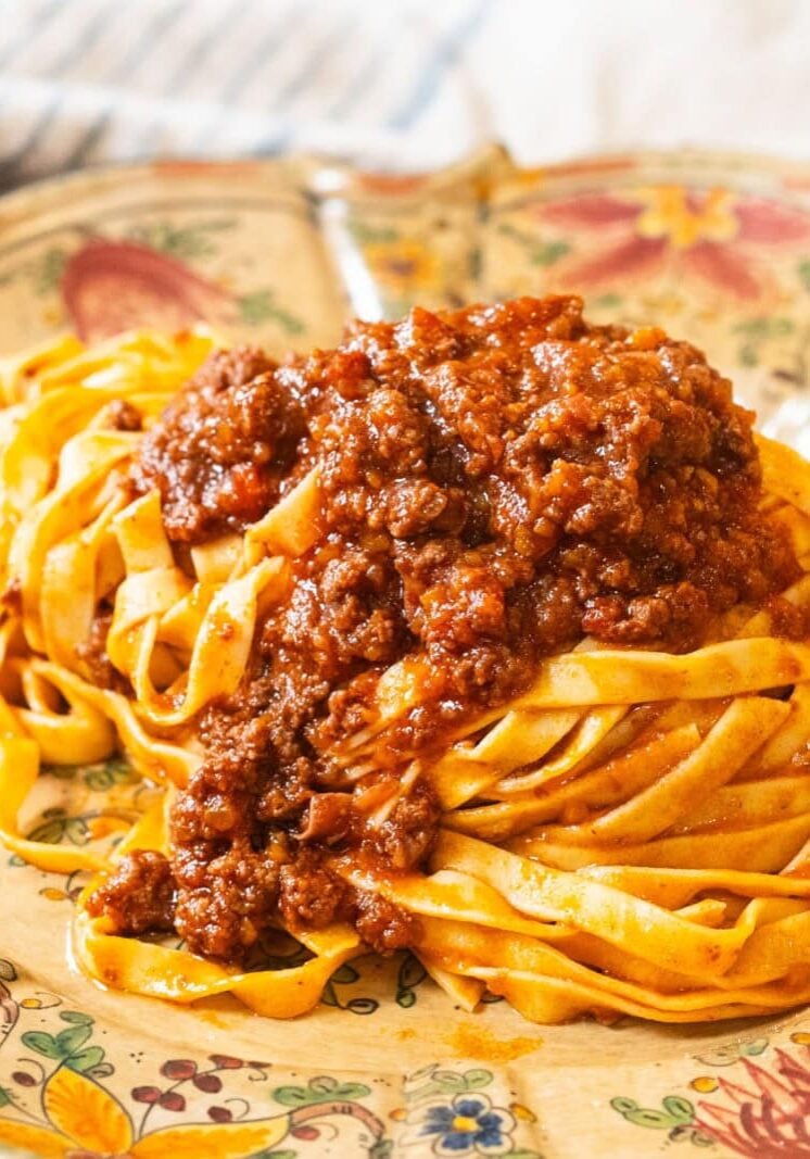 A plate of tagliatelle pasta topped with meat ragù sauce, served on a decorative floral dish with a cloth in the background.
