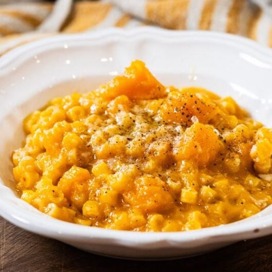 A white bowl filled with creamy pumpkin risotto, garnished with black pepper, sits on a wooden surface with a striped cloth in the background.