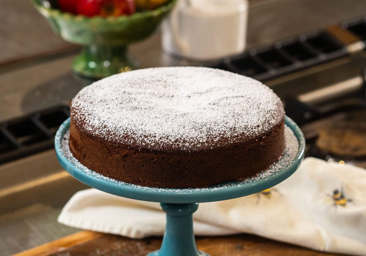 A round chocolate cake dusted with powdered sugar sits on a blue cake stand, with a white cloth and fruit bowl in the background.