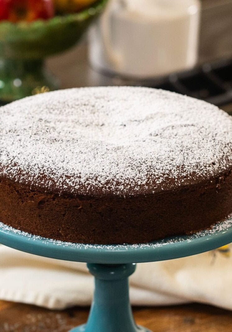 A round chocolate cake dusted with powdered sugar sits on a blue cake stand, with a white cloth and fruit bowl in the background.