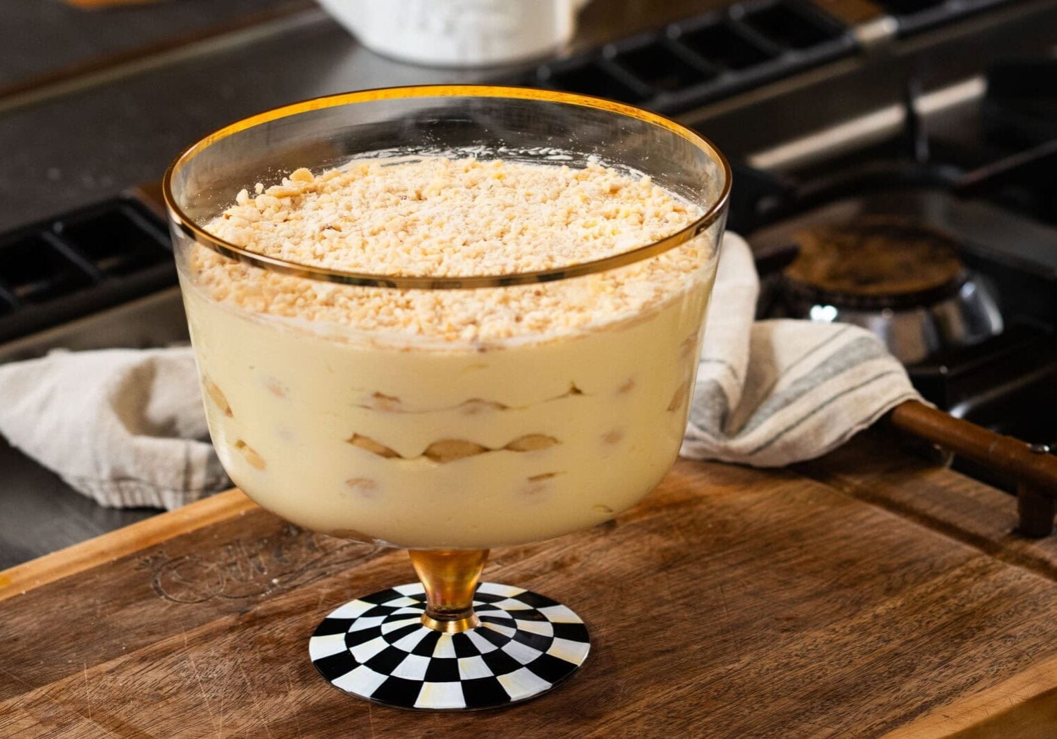 A glass trifle dish filled with layers of creamy pudding, bananas, and crumbled cookies sits on a wooden countertop in a kitchen.