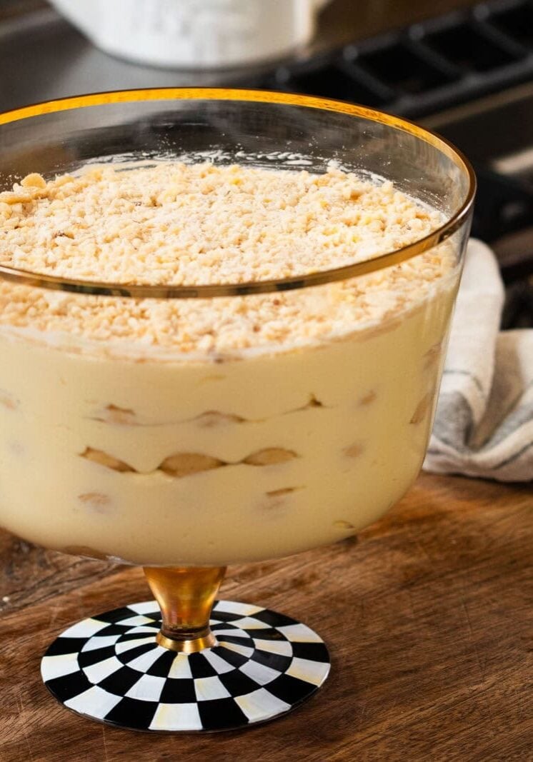 A glass trifle dish filled with layers of creamy pudding, bananas, and crumbled cookies sits on a wooden countertop in a kitchen.