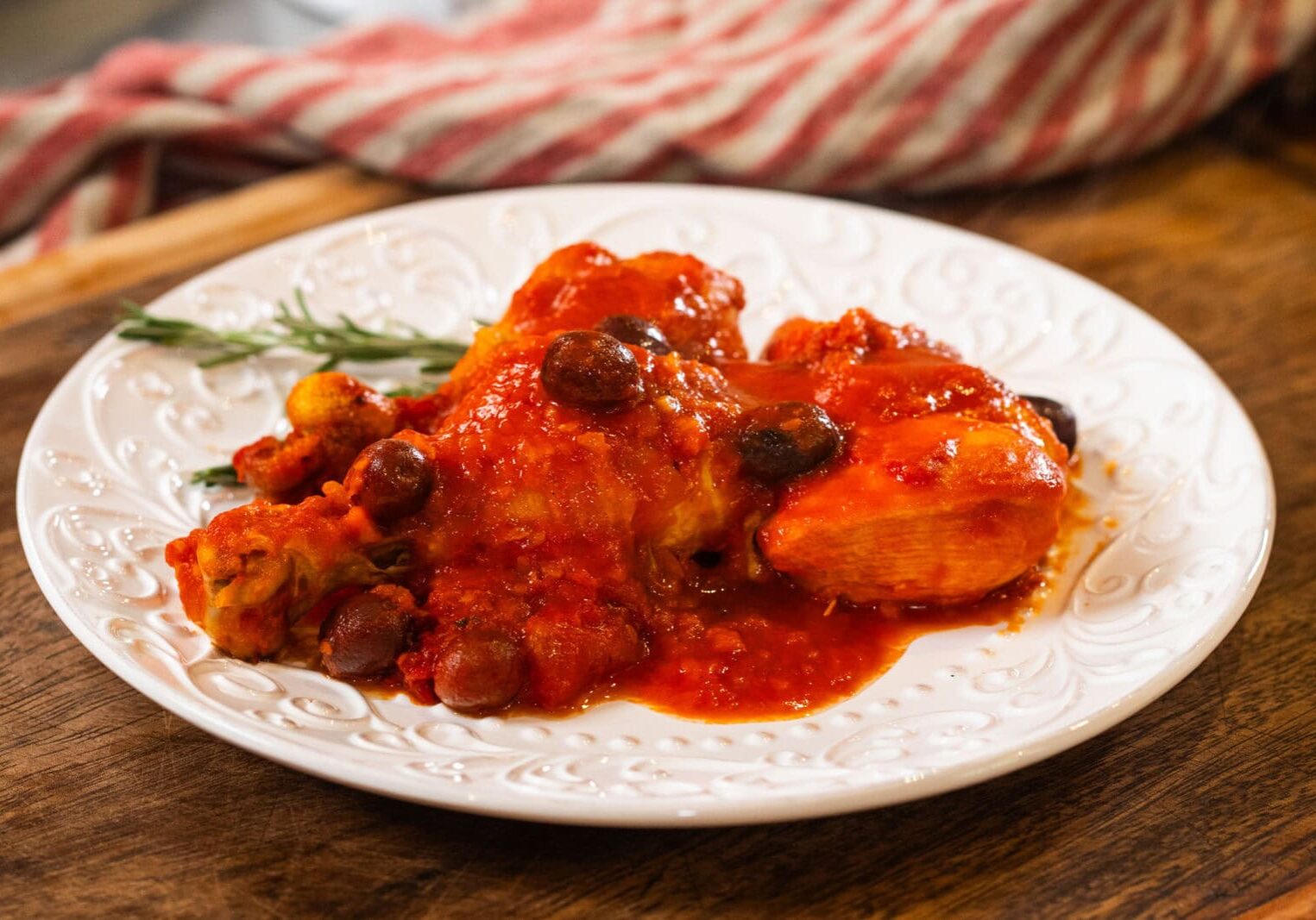 A white plate with chicken in red tomato sauce, garnished with olives and herbs, sits on a wooden surface with a striped cloth in the background.