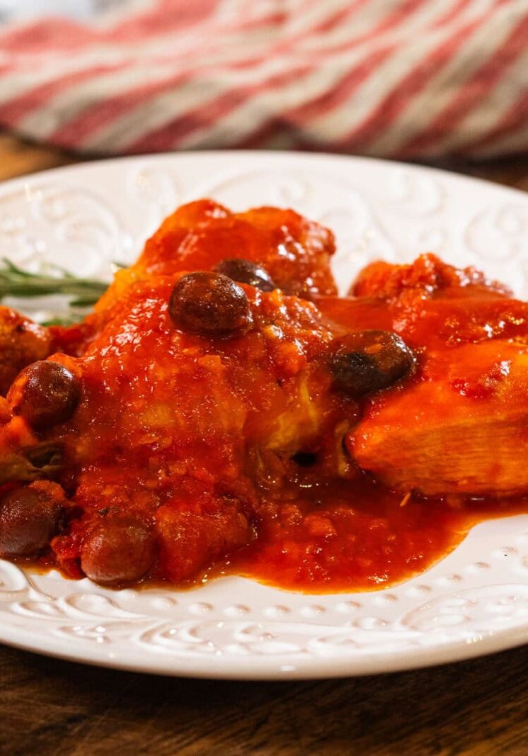 A white plate with chicken in red tomato sauce, garnished with olives and herbs, sits on a wooden surface with a striped cloth in the background.