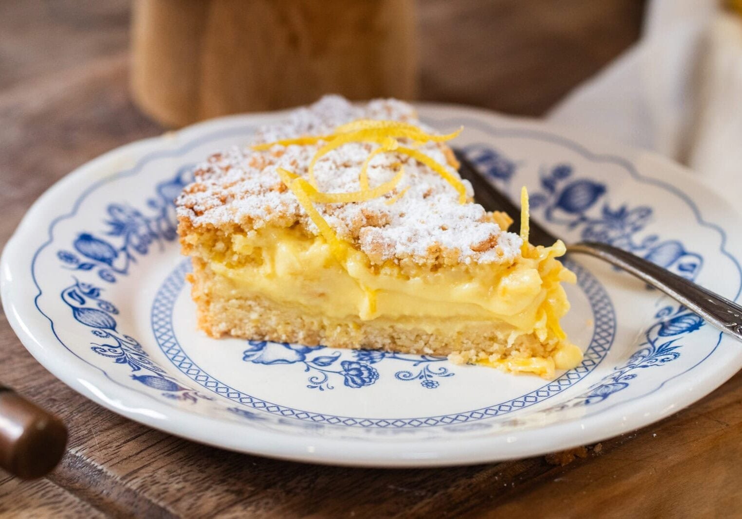 A slice of lemon cream pie with powdered sugar and lemon zest on top, served on a blue and white plate with a fork.