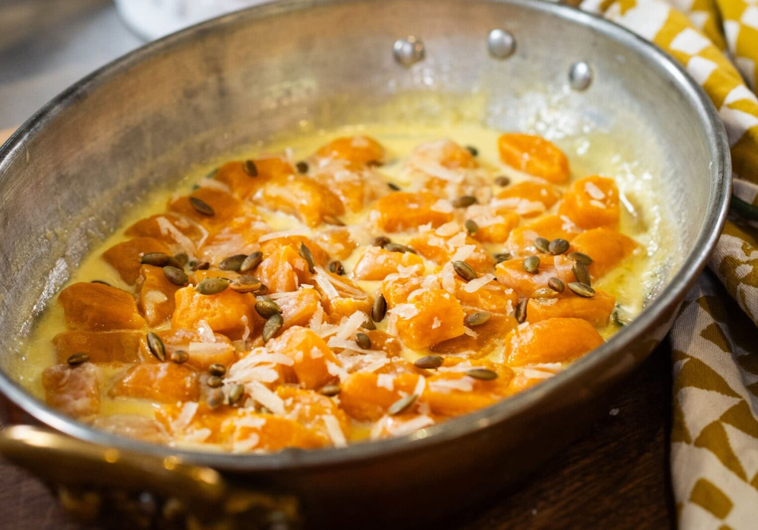 A metal pan contains cooked gnocchi in a creamy sauce, topped with grated cheese and pumpkin seeds.